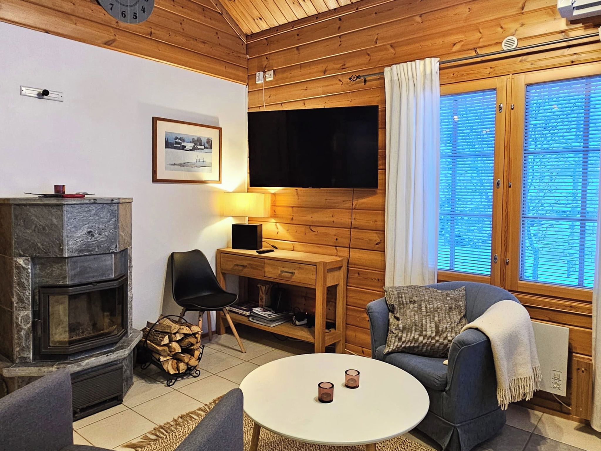 Un intérieur de chalet chaleureux avec une cheminée, un bureau avec un ordinateur, un canapé confortable et une grande fenêtre avec des stores.