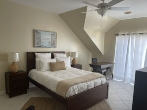 A bedroom with a bed, two bedside tables, a desk, a chair, a ceiling fan, and a painting on the wall. - Lagoon Luxury at Sandyport (Nassau)