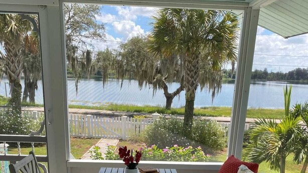 Une terrasse couverte avec une table et des chaises, un banc rembourré et une vue sur un lac bordé d’arbres.