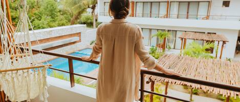 A person standing on a balcony overlooking a pool with a thatched roof structure and lush greenery in the background.