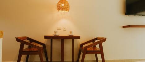 A room with a wooden table and two chairs, a hanging woven light fixture, a wall-mounted television, and a power outlet.