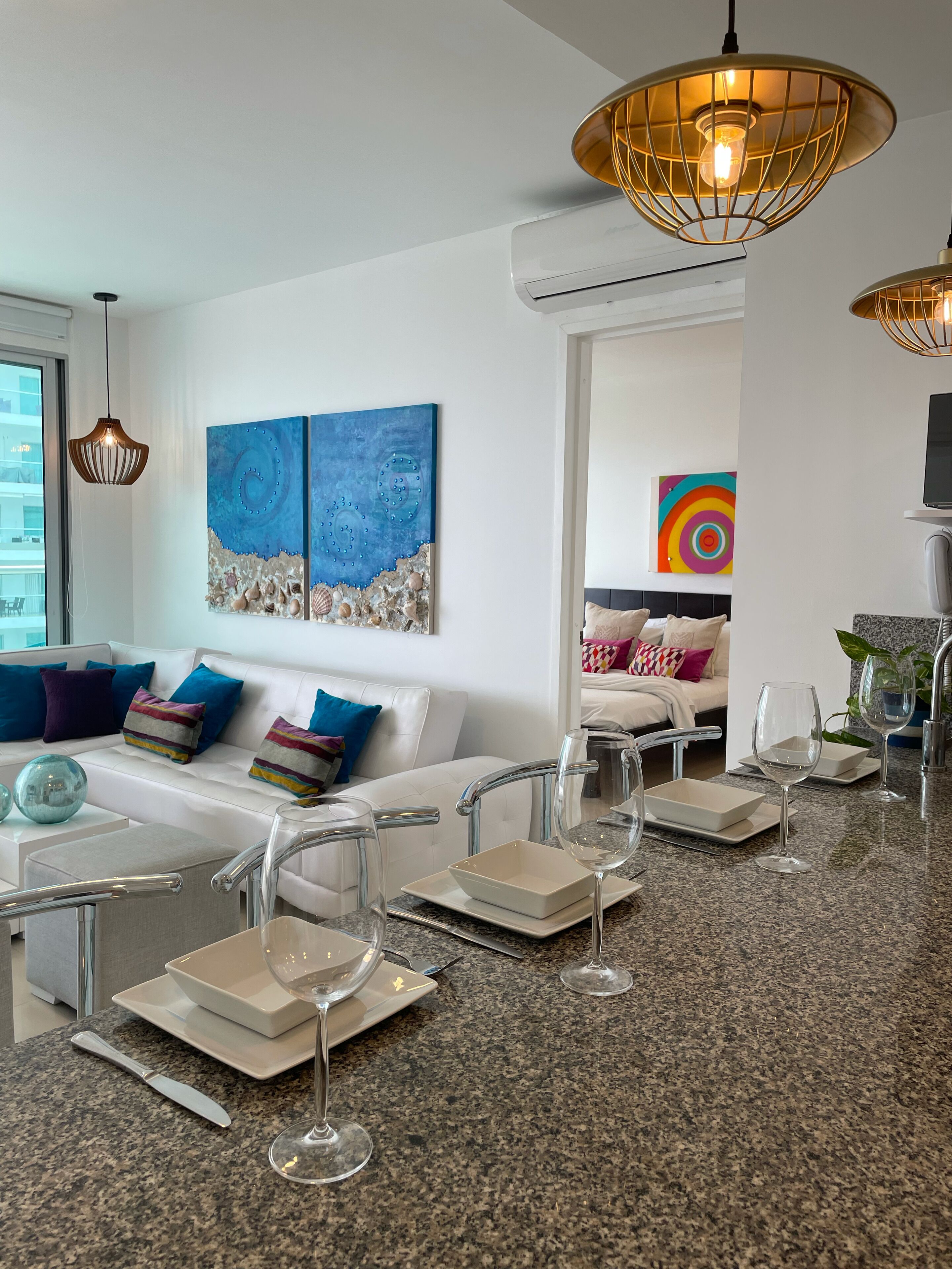 A modern kitchen with granite countertops, white cabinetry, and a central island. There are two pendant lights, a large window, and a colorful abstract painting on the wall.