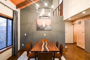 A dining area with a wooden table and chairs, a modern chandelier, and a painting on the wall.