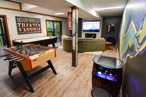 A room with a foosball table, a pool table, a sofa, a television, and a mural.