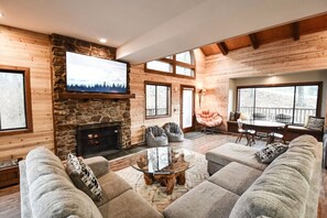 A cozy living room with a stone fireplace, a flat-screen TV, and a large window overlooking a deck.