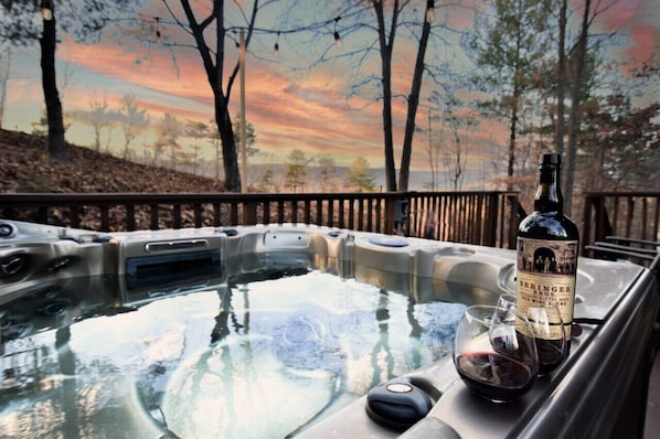 A hot tub with a bottle of wine and glasses on a car's roof, surrounded by trees and a sunset.