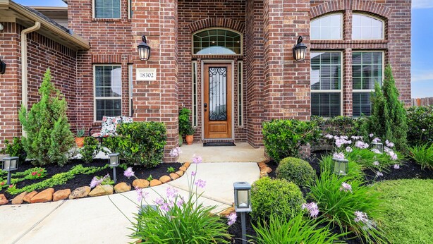 A brick house with a front door, number 18302, and a well-maintained garden with various plants and flowers. - Elegant spacious home with water view. Prime location- Richmond, Katy, Houston, Rosenberg. (Richmond)