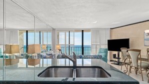 A modern kitchen with a stainless steel sink, a dining area with a TV, and a view of the ocean through large windows.