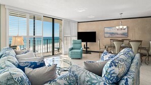 A spacious living room with a large sofa, a small table, and a chair. There is a balcony with a view of the beach.