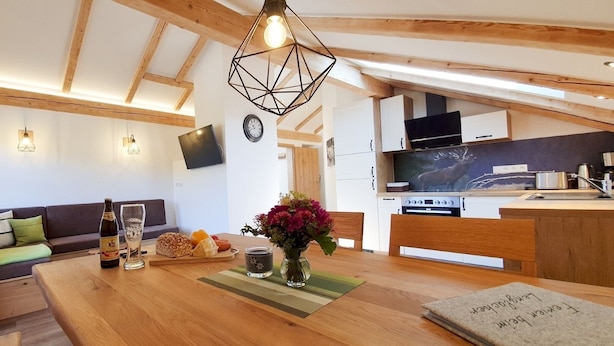 A cozy living room with a wooden ceiling, a dining table with a vase of flowers, a sofa with cushions, and a kitchen area with a TV and modern appliances. - Vacation apartment Schwalbennest (Chieming)