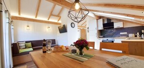 Uma sala de estar acolhedora com teto de madeira, uma mesa de jantar com um vaso de flores, um sofá com almofadas e uma área de cozinha com TV e eletrodomésticos modernos.