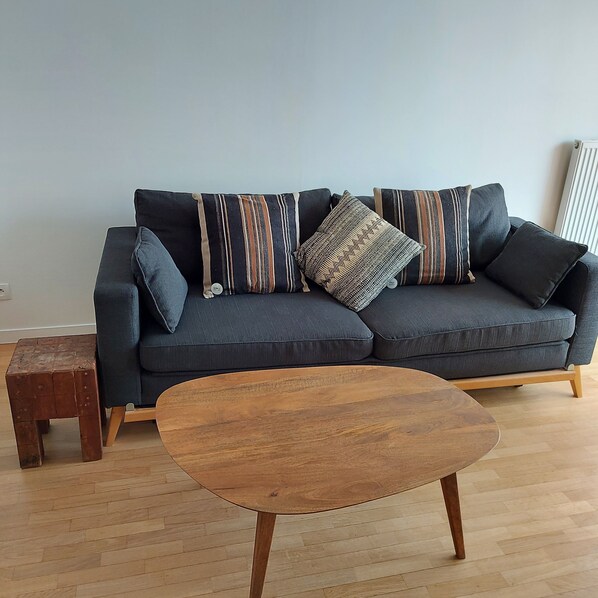 A dark grey sectional sofa with multiple pillows, a small wooden stool, and a round wooden coffee table in a living room with wooden flooring.