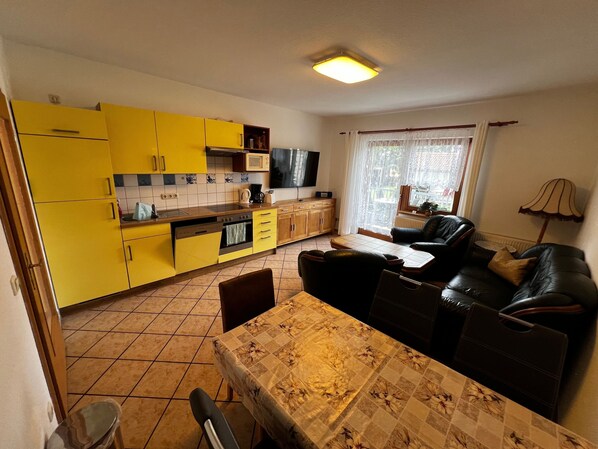 A compact living space with a kitchen featuring yellow cabinets, a dining area with a table and chairs, a black leather sofa, and a window with curtains. - Vacation apartment 2 - Wiedemann Heiko (Dargen)