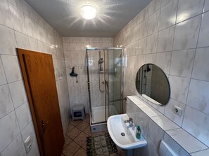 Casa de banho com chuveiro de vidro, pia com espelho e porta para outro espaço.