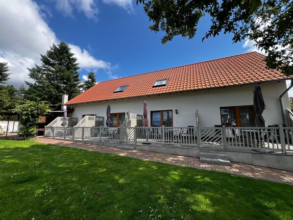 En bygning med to etager, rødt tegltag, en overdækket terrasse og et hvidt hegn.