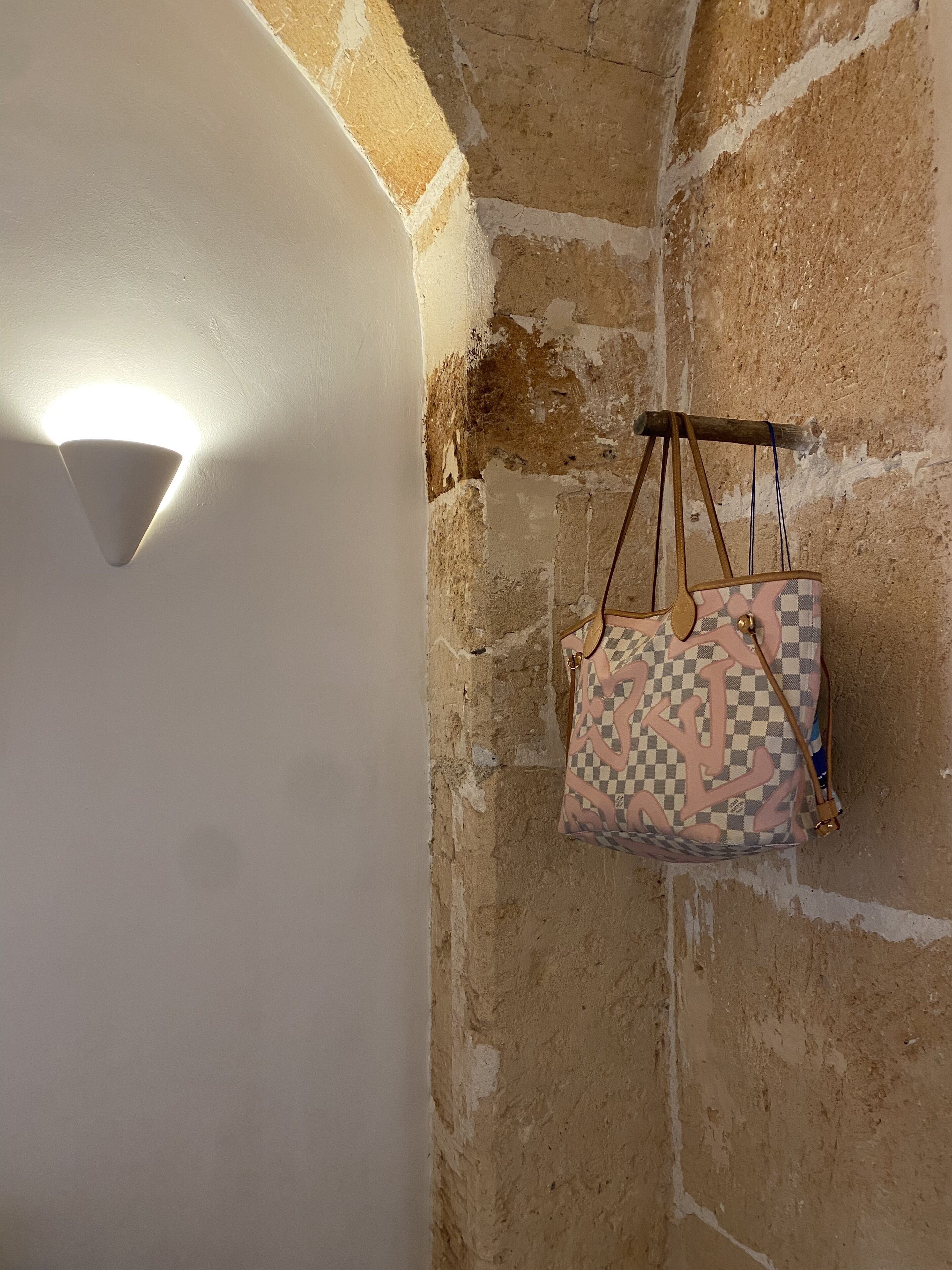 Eine Ecke eines Raumes mit weißer Wand und freigelegtem Mauerwerk. Eine Pendelleuchte ist sichtbar, und eine gemusterte Tasche hängt von der Decke.