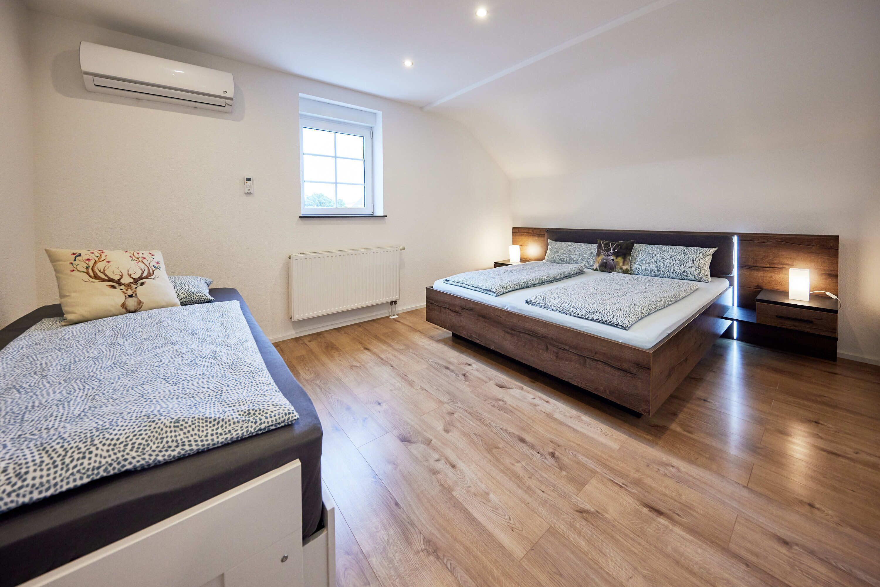 A bedroom with a large bed, a smaller bed, a window, an air conditioner, and a bedside table with a lamp.