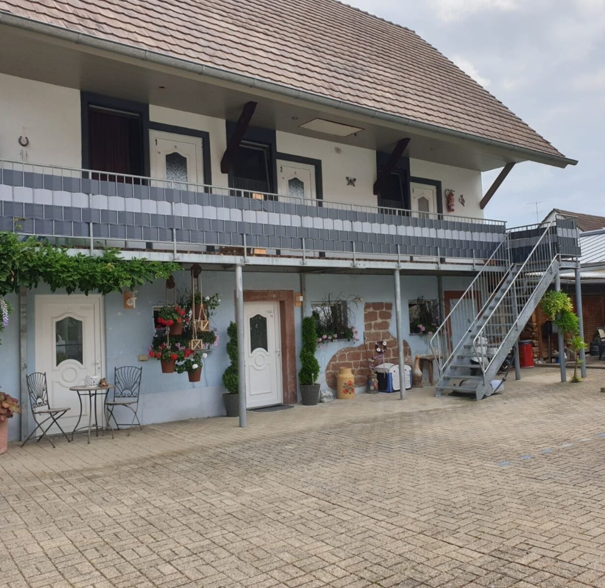 Un bâtiment à deux étages, doté d’un toit recouvert de tuiles, d’un balcon avec des rampes en métal et d’un escalier menant à l’étage supérieur. Au rez-de-chaussée, on trouve des plantes en pot et du mobilier d’extérieur.