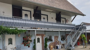 A two-story building with a tiled roof, a balcony with metal railings, and a staircase leading to the upper floor. There are potted plants and outdoor furniture on the ground floor.