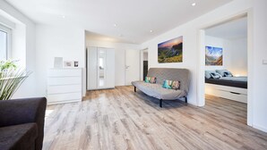Een moderne woonkamer met een houten vloer, een grijze bank met kleurrijke kussens en een bed met een blauw hoofdeinde.