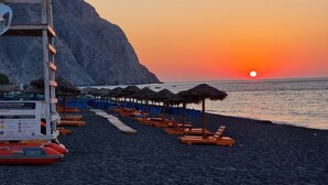 Una playa con sillones de madera y sombrillas de paja, una torre de salvavidas y un atardecer sobre el océano.