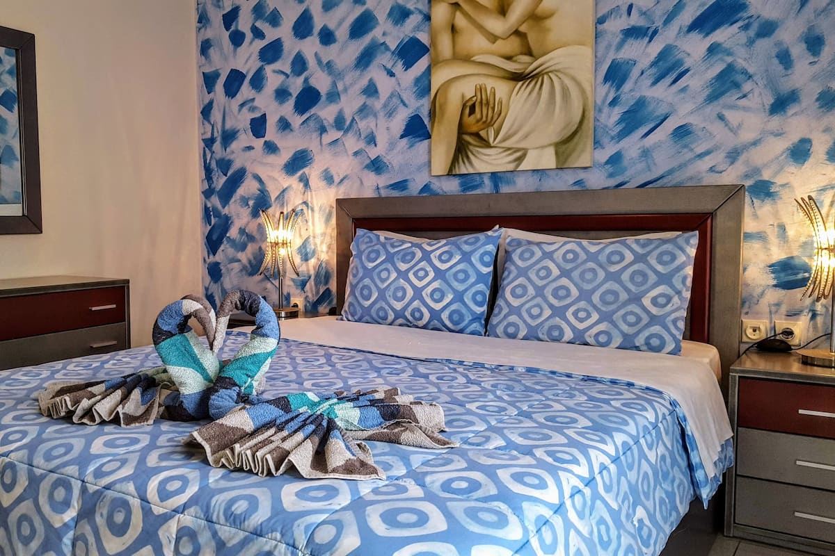 Un dormitorio con una colcha estampada, cabecera, mesitas de noche con lámparas y obras de arte en las paredes.