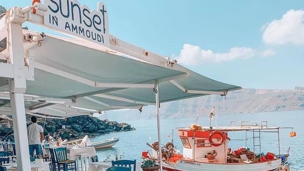 Un restaurante frente al mar con terraza al aire libre, un barco atracado cerca y vistas claras de la costa rocosa.