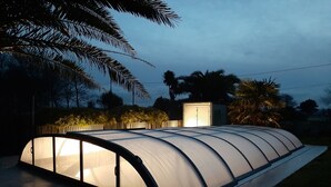 A greenhouse structure with a curved roof and transparent walls, surrounded by palm trees and a small shed in the background.