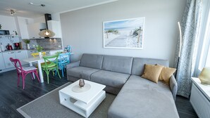 En moderne stue med en grå sofa, et hvidt sofabord og farverige stole. Der er et køkkenområde med hvide skabe og et spisebord med blå stole. Et strandmotiv er vist på væggen.