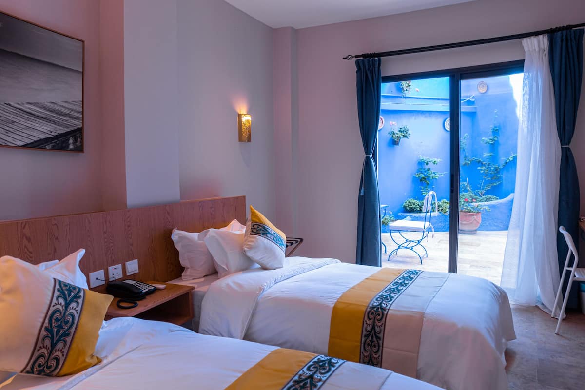 A hotel room with two beds, a small table, a chair, and a view of the outside.
