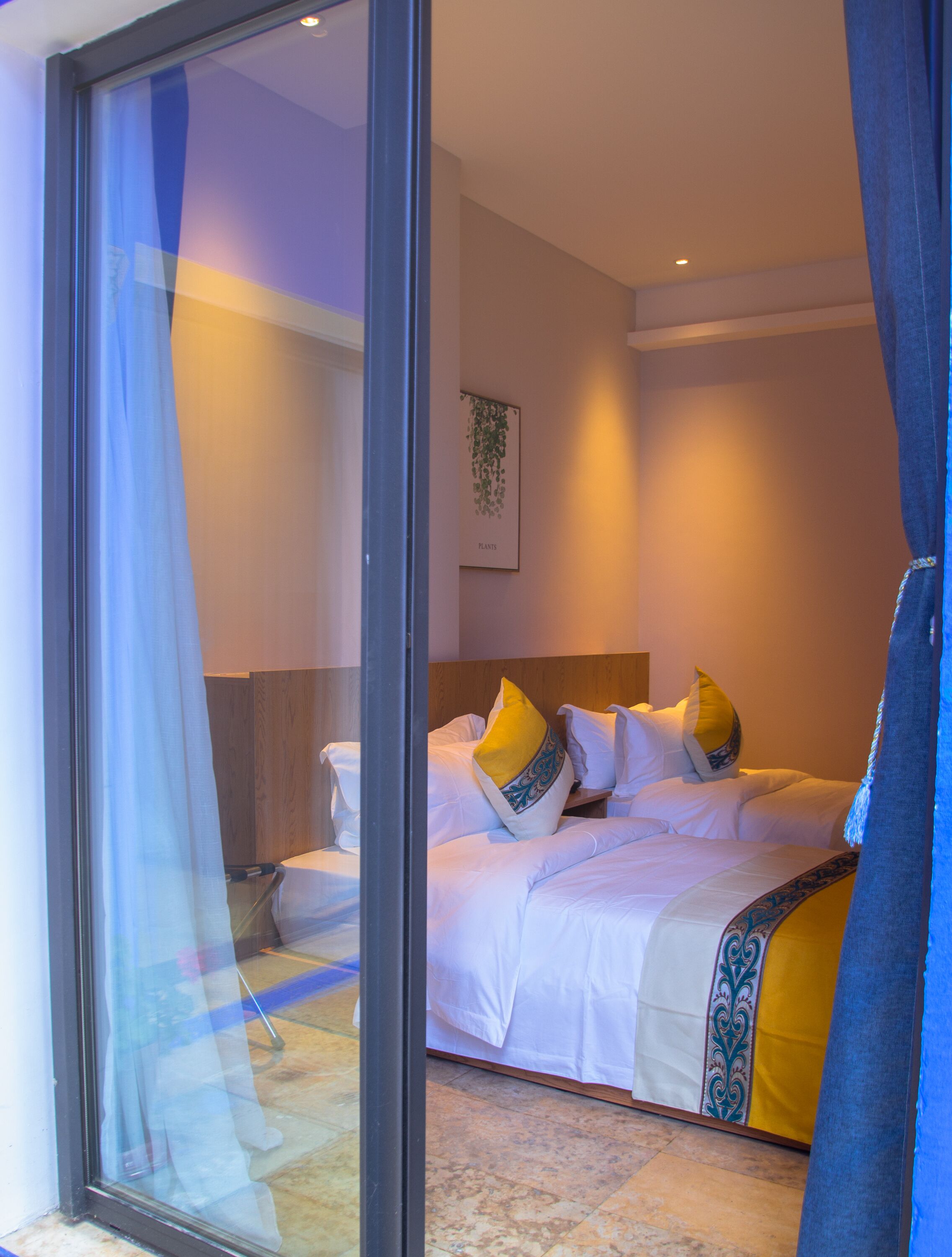 Habitación de hotel con cama, ventana con cortinas translúcidas, planta en la pared y cabecero de madera.