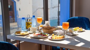 A breakfast table with various food items and drinks, a wicker basket, and a view of a building through a window. - VANCII HOTEL (Chefchaouen)