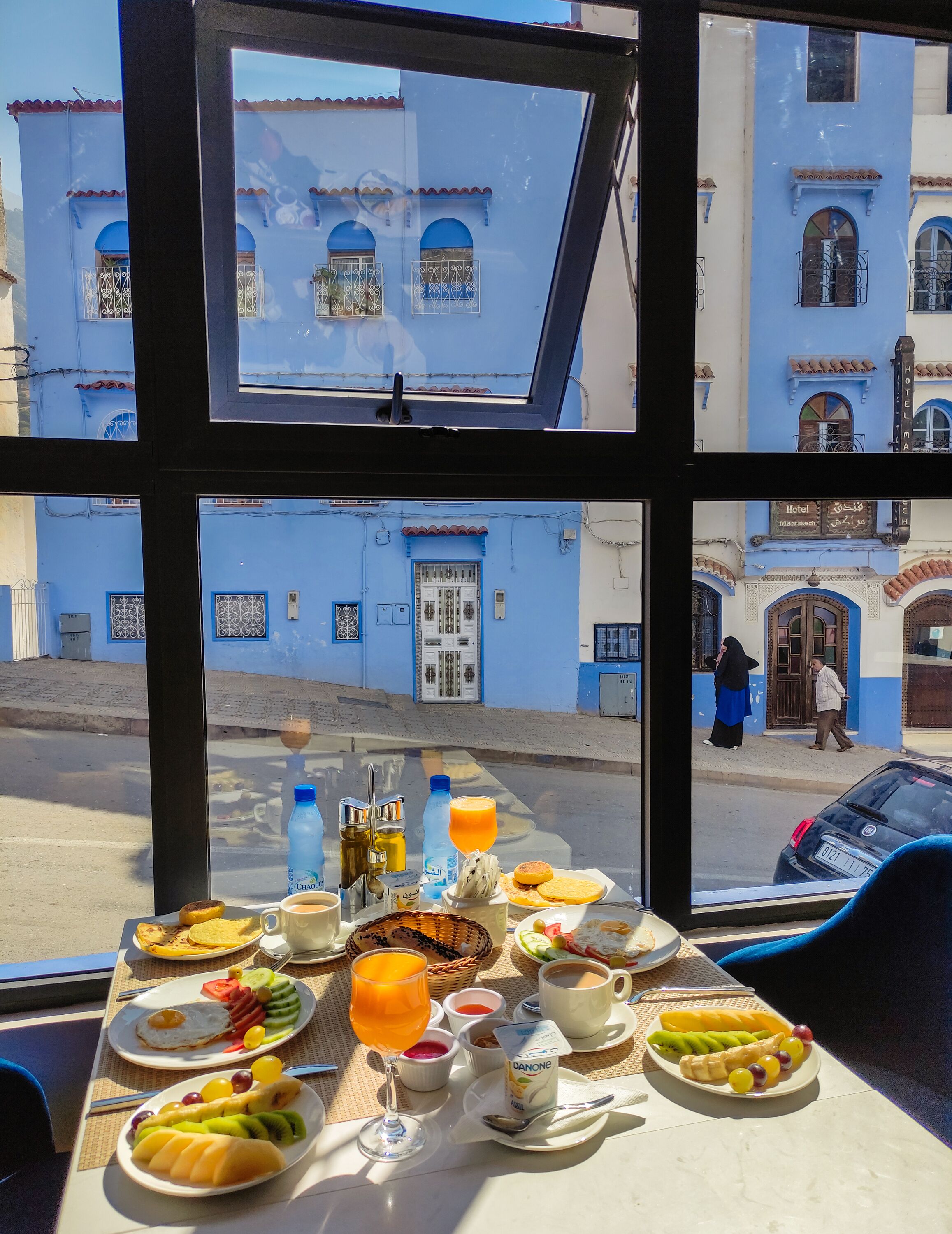 A breakfast table with various food items and drinks, a window view of a street with buildings, and a car parked outside.