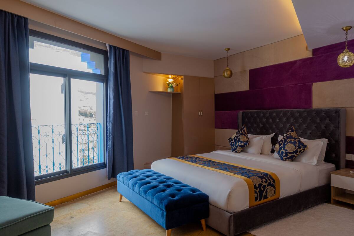 A modern hotel room with a large bed, a bench, a small table, and a view of the outside.