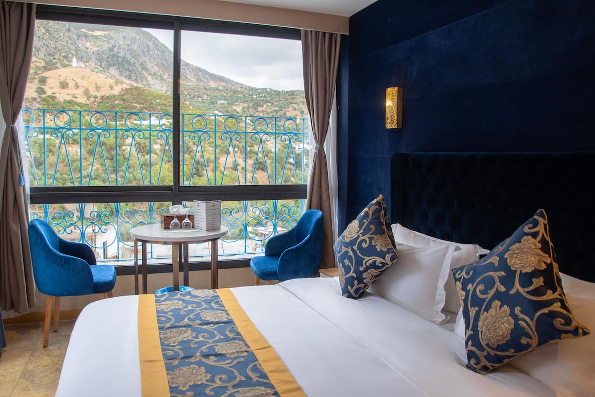 A hotel room with a large bed, two blue chairs, a small table with a bottle and glasses, and a view of a mountainous landscape through a window with blue iron railings.