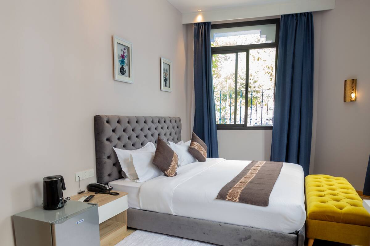 A modern hotel room with a large bed, a bedside table with a phone and a coffee maker, a small desk, a yellow ottoman, and a window with blue curtains.