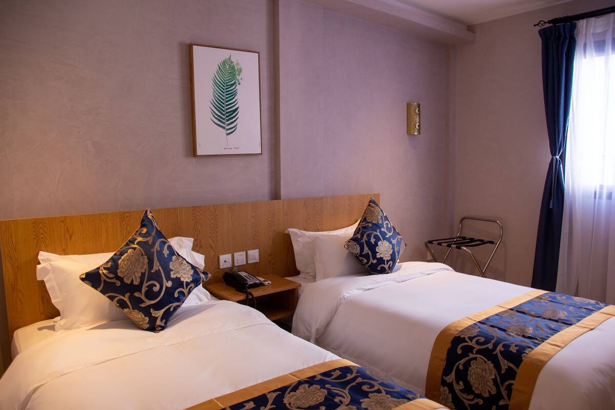 A hotel room with two beds, a wooden headboard, a framed botanical print on the wall, a telephone on the nightstand, and a window with curtains.