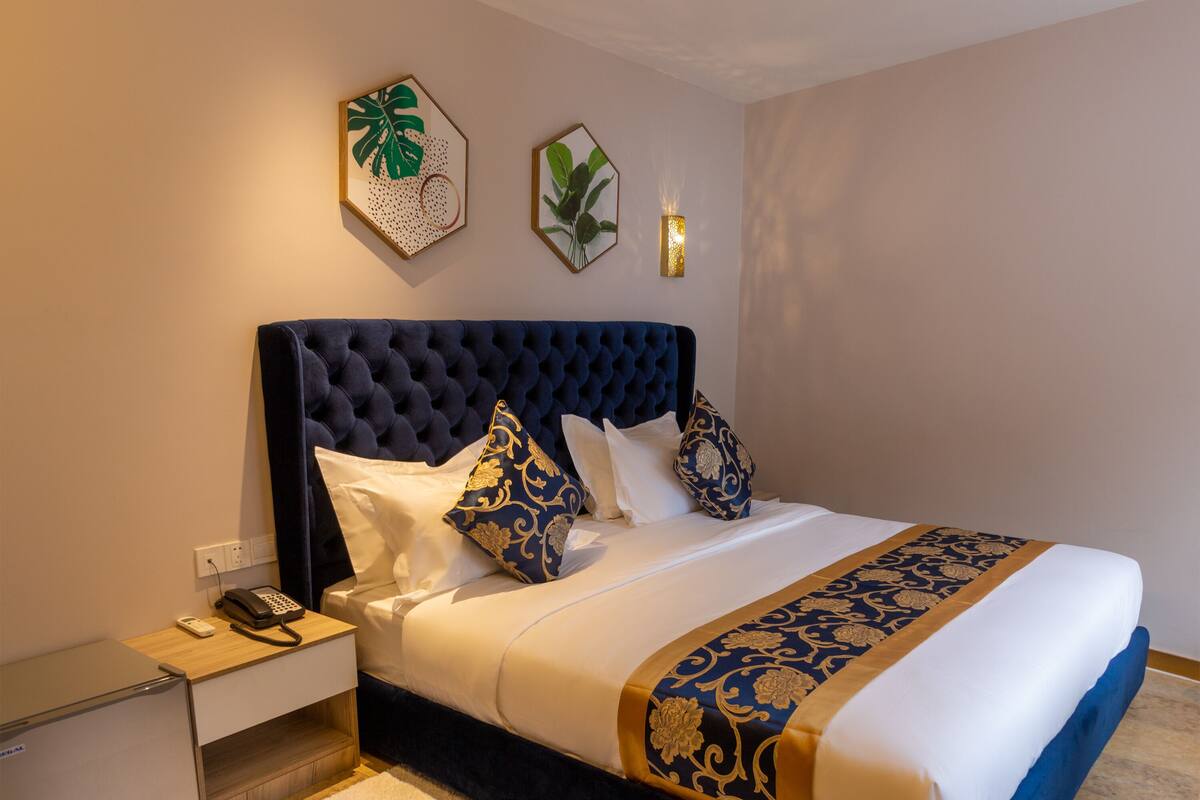 A neatly made bed with a dark headboard, white bedding, and decorative pillows. There is a bedside table with a phone and a lamp.