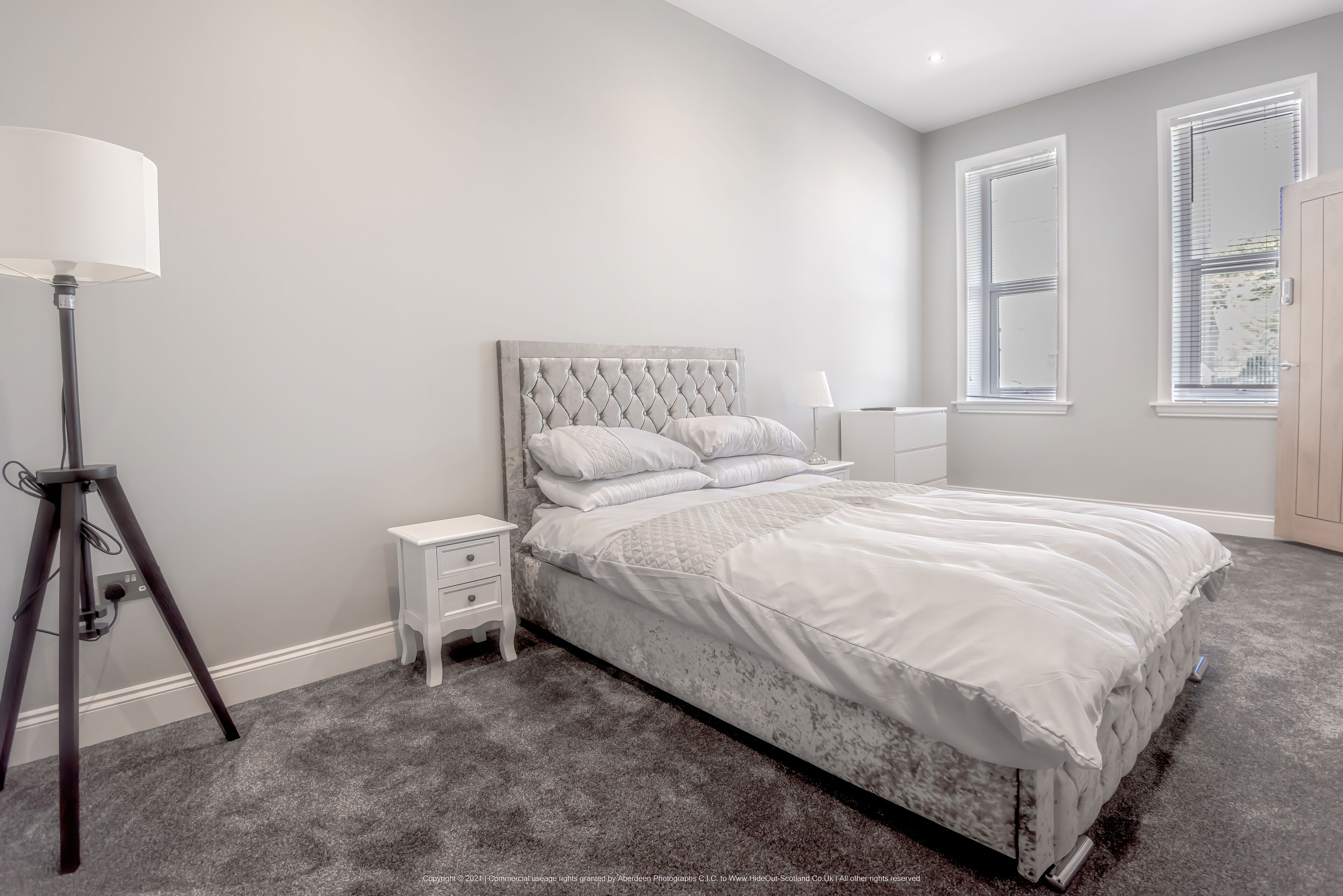 A bedroom with a tufted headboard, a white bed, a bedside table, a lamp, and a window with blinds.