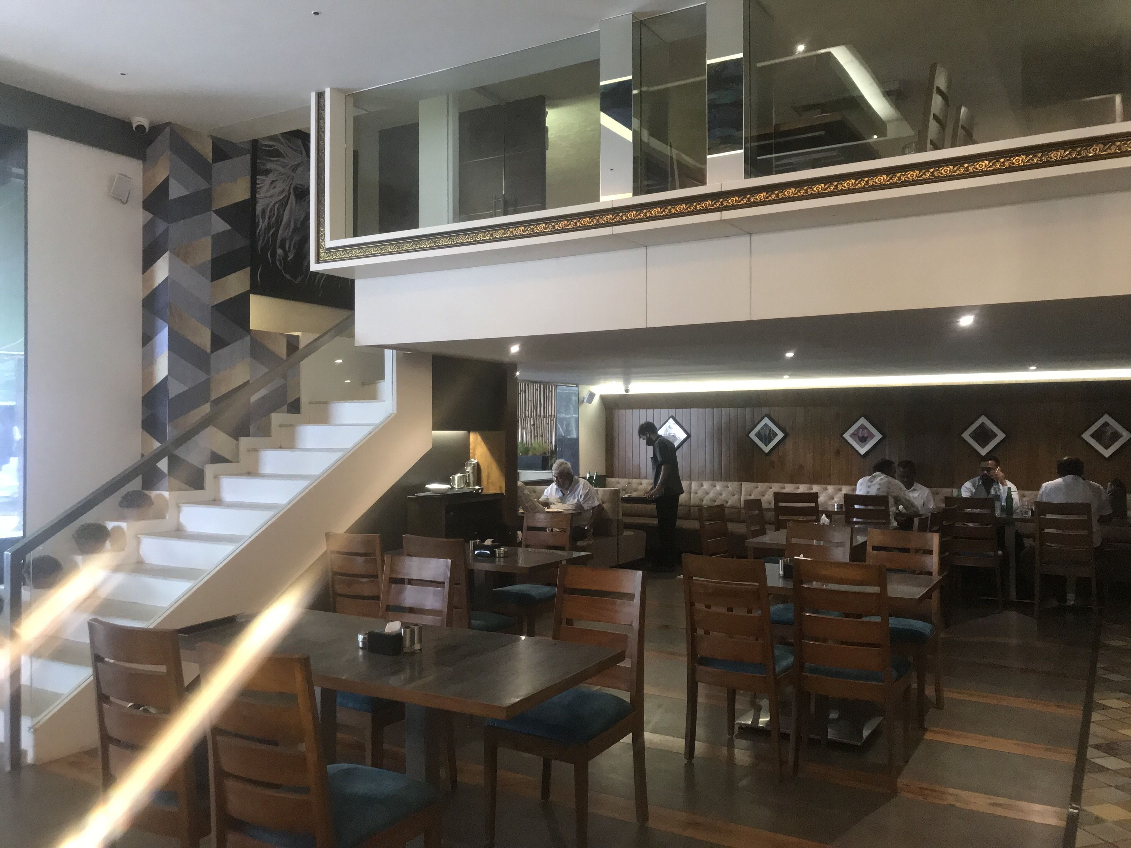 A modern restaurant with a staircase leading to an upper level, wooden tables and chairs, and a patterned wall.
