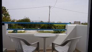 A balcony with two white plastic chairs and a small round table. A blue railing separates the balcony from the greenery below.