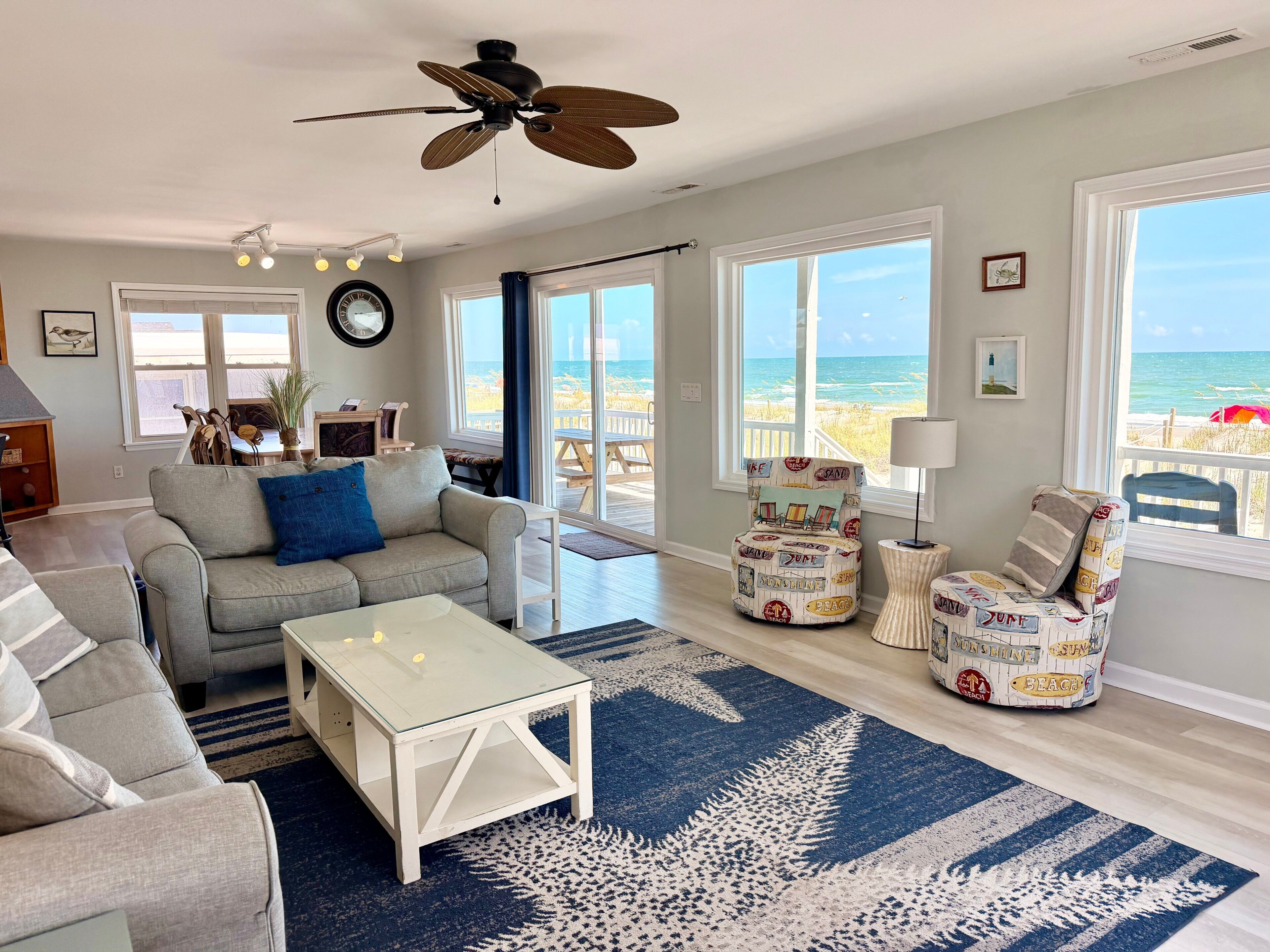 A living room with a sofa, coffee table, and chairs, large windows with a view of the beach, and a ceiling fan.