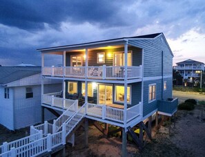 A two-story beachfront house with a balcony, a staircase, and a view of the ocean.