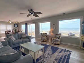 A living room with a sofa, coffee table, and two patterned chairs. There is a ceiling fan, a clock, and a view of the ocean through large windows.