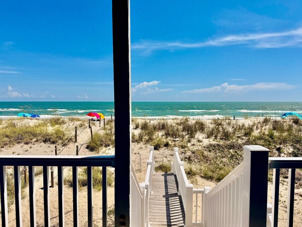 A wooden deck leading to a beach with dunes and a clear blue sky.