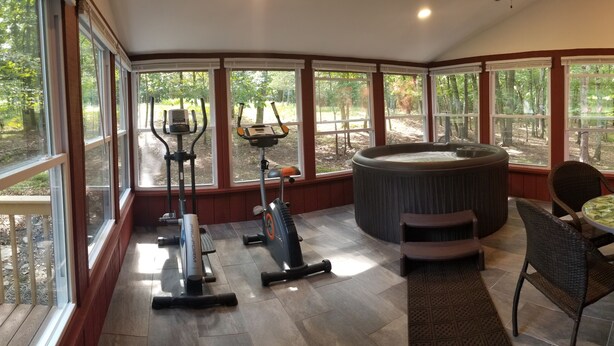 Ein Wintergarten mit Trainingsgeräten, einer Whirlwanne und einem Essbereich mit Blick auf den Wald.