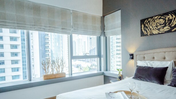 A hotel room with a large bed, a window with blinds, a view of tall buildings, and a decorative wall piece.