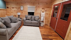 A cozy room with two grey sofas, a white rug, wooden walls, a red door, and a window with blinds. - Beech View Haven- Custom Log Home in Hocking Hills (Logan)