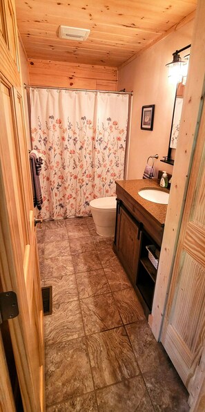 A bathroom with a wooden ceiling, a shower curtain with floral patterns, a toilet, a sink with a mirror, and a towel rack. - Beech View Haven- Custom Log Home in Hocking Hills (Logan)