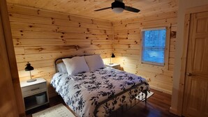 A wooden cabin bedroom with a bed, bedside tables, a ceiling fan, and a window with blinds. - Beech View Haven- Custom Log Home in Hocking Hills (Logan)